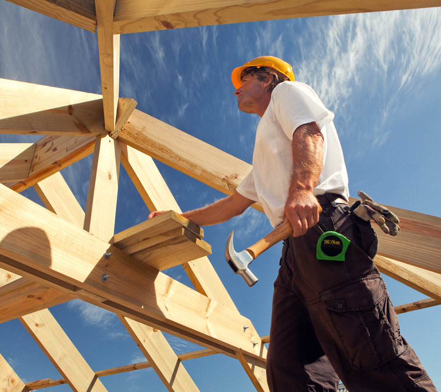 Bauarbeiter mit Hammer, steht im Freien unter Dachbalken aus Holz.
