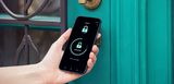 Eine Hand hält ein Smartphone vor eine elegant-antik gestaltete blaugrüne Haustür. Auf dem Smartphone ist eine Smart-Home-App geöffnet, die offenbar die Haustür entriegelt.Das Bild vermittelt den Einsatz smarter Sicherheitstechnik zur Zugangskontrolle.