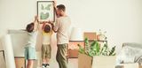 Ein Vater hängt mit zwei Kindern ein Bild im neuen Zuhause auf – Umzugskartons und Pflanzen stehen noch ungeordnet herum. Dank staatlicher Förderung wird der Wohntraum für Familien Realität.