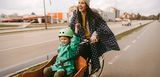 Eine junge Frau fährt auf einem Lastenfahrrad mit einem Kind mit Fahrradhelm im Laderaum. Beide lachen und haben Regenmäntel an. Sie fahren auf einem von der Autostraße abgegrenzten Radweg.