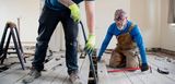 Zwei Heimwerker sanieren in Eigenregie den Holzboden eines Hauses. Das Bild zeigt Renovierungsarbeiten mit Eigenleistung und steht für den passenden Versicherungsschutz bei einer Haussanierung.