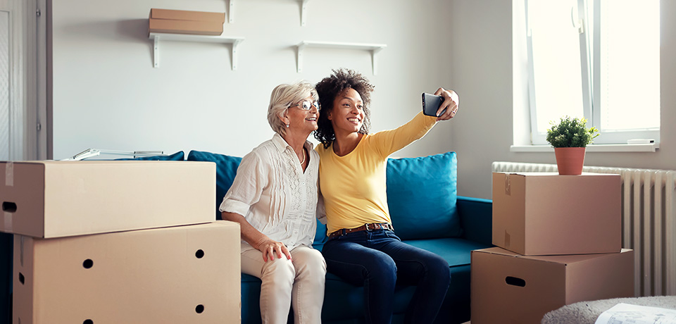 Eine Mutter hat ihrer Tochter beim Auszug geholfen. Überall stehen Kartons herum. Zur Erinnerung machen sie eine Foto in der neuen Wohnung. 