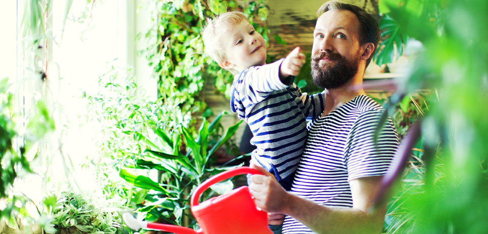 Vater mit Kind auf dem Arm und roter Gießkanne umgeben von Zimmerpflanzen