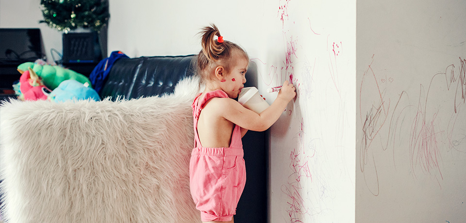 Kleinkind kritzelt mit Filzstift an eine weiße Wand im Wohnzimmer