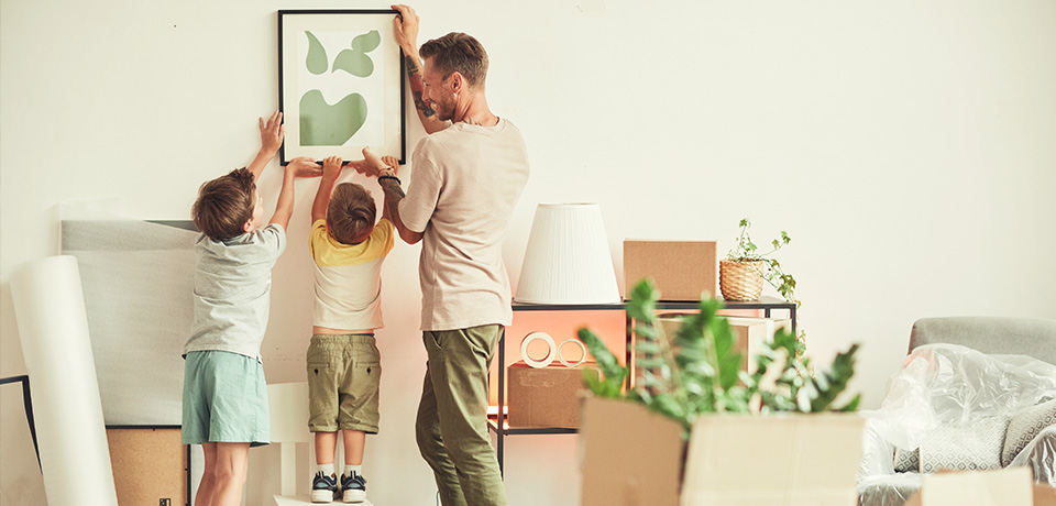 Familie richtet neues Heim ein – mit KfW-Förderung zum Eigenheimglück Ein Vater hängt mit zwei Kindern ein Bild im neuen Zuhause auf – Umzugskartons und Pflanzen stehen noch ungeordnet herum. Dank staatlicher Förderung wird der Wohntraum für Familien Realität.