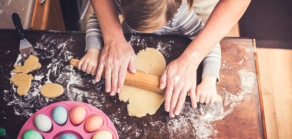 Mutter und Kleinkind machen Osterplätzen. Sie rollen den Teig aus, stechen Osterhasen mit Formen aus. Die Ostereier sind schon gefärbt. 