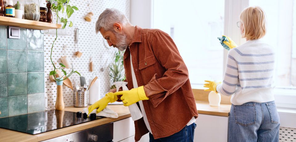 Mann mit graumeliertem Haar und gelben Gummihandschuhen reinigt den Herd, während im Hintergrund eine Frau mit gelben Gummihandschuhen das Fenster putzt.