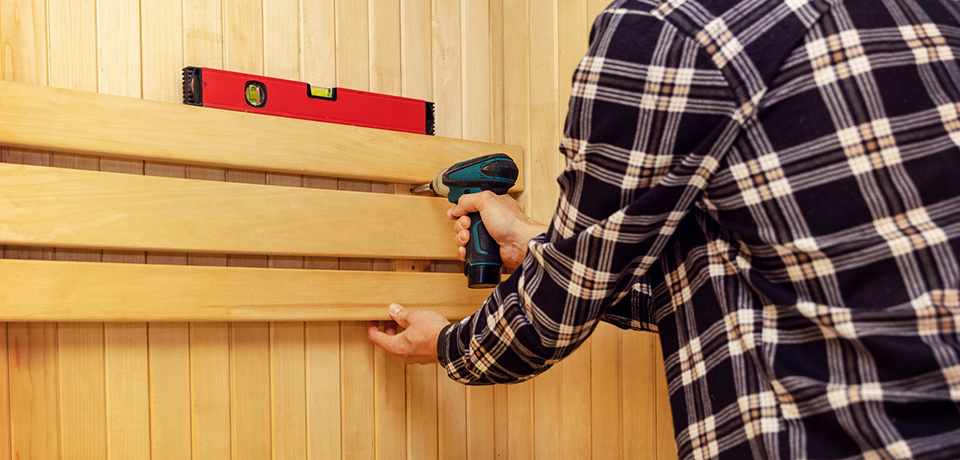 Person beim Bau einer Heimsauna: Holzverkleidung wird mit einer Bohrmaschine an der Wand befestigt und mithilfe einer Wasserwaage exakt ausgerichtet.