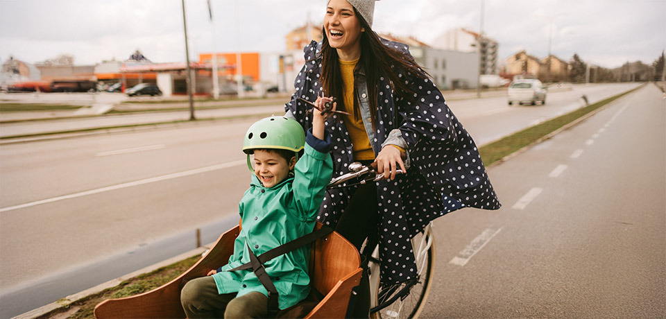 Richtig versichert mit dem Lastenrad unterwegs - so macht es Spaß! Eine junge Frau fährt auf einem Lastenfahrrad mit einem Kind mit Fahrradhelm im Laderaum. Beide lachen und haben Regenmäntel an. Sie fahren auf einem von der Autostraße abgegrenzten Radweg.