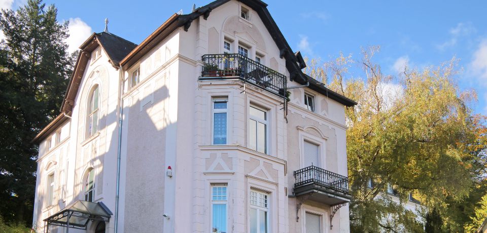 Außenansicht eines mehrstöckigen, hellen Wohngebäudes mit Ornamentfassade, Balkonen und umliegenden Bäumen bei sonnigem Wetter.