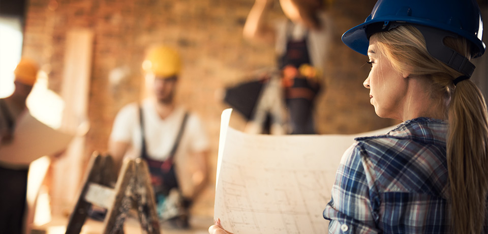 Eine Architektin fungiert als Bauleiterin und studiert auf der Baustelle den Bauplan. Im Hintergrund sieht man die Bauarbeiter. 