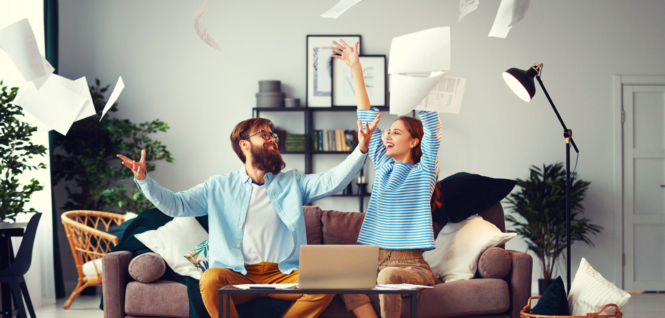 Junges Paar auf moderner Couch werfen Papiere in die Luft und freuen sich. So viele Dokumente müssen nicht oder nicht in Papierform aufbewahrt werden.  