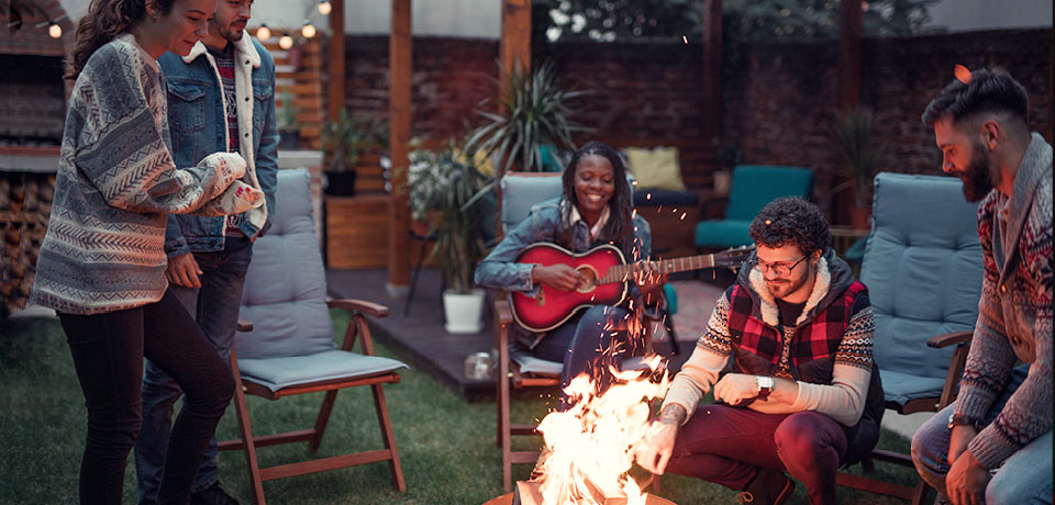 Junge Leute feiern im Garten um eine Feuerschale herum. Ein Mädchen spielt Gitarre. Vermutlich singen sie gemeinsam.