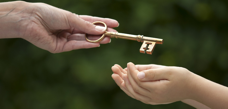 Ein großer goldener Schlüssel wird von einer älteren Frauenhand an eine Kinderhand gereicht. 