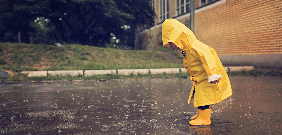 Kleines Kind in gelben Gummistiefeln und gelber Regenjacke steht im Regen in einer Pfütze. Das Gesicht ist verborgen unter der gelben Kapuze, aber die Körperhaltung zeigt Neugierde und Interesse. 
